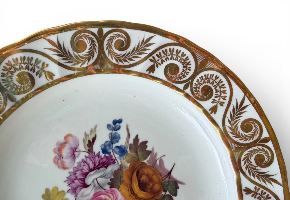 A circular white porcelain dish featuring a hand-painted floral centrepiece framed by a gilded scrollwork rim. View II