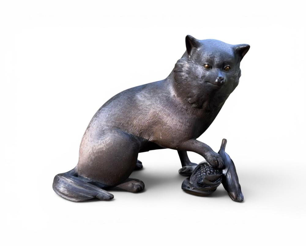 A Japanese bronze sculpture depicting a seated tanuki resting its paw on corn husks, featuring textured fur engraving and a dark patinated finish.