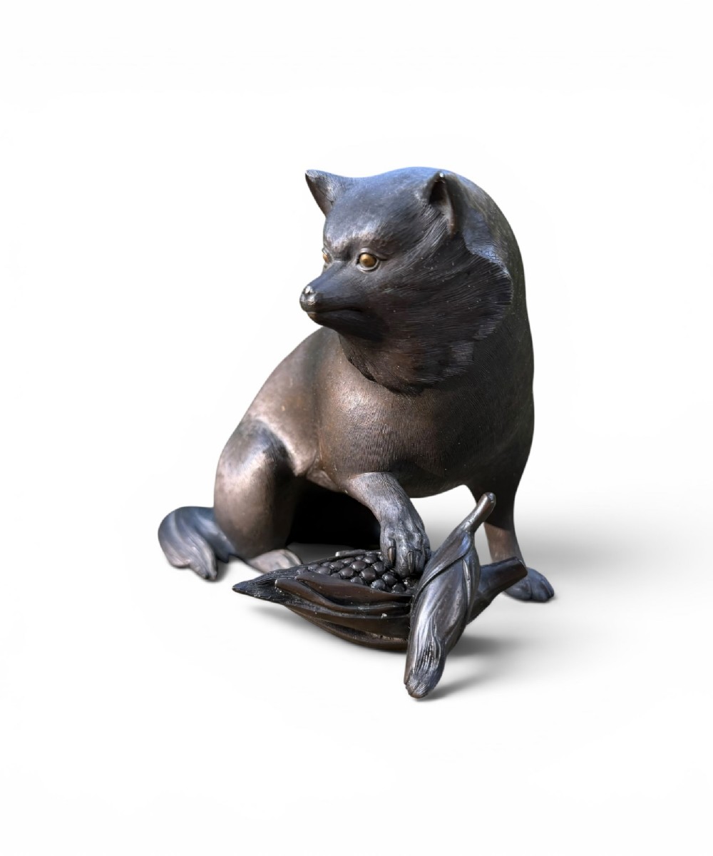 A Japanese bronze sculpture depicting a seated tanuki resting its paw on corn husks, featuring textured fur engraving and a dark patinated finish. View II