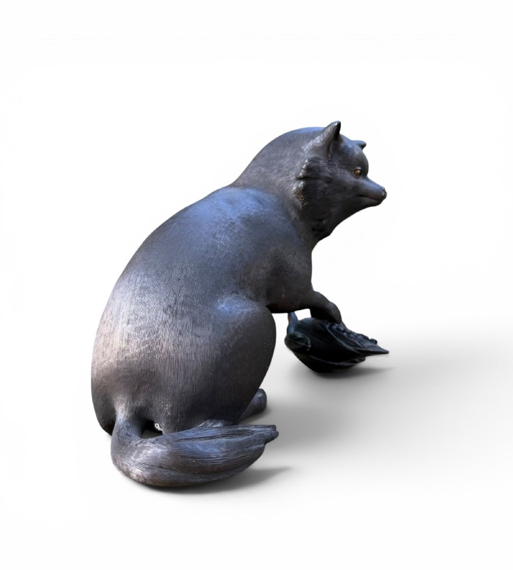 A Japanese bronze sculpture depicting a seated tanuki resting its paw on corn husks, featuring textured fur engraving and a dark patinated finish. View IV