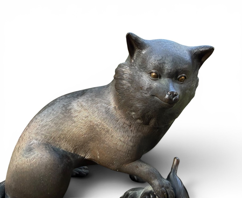 A Japanese bronze sculpture depicting a seated tanuki resting its paw on corn husks, featuring textured fur engraving and a dark patinated finish. View VI