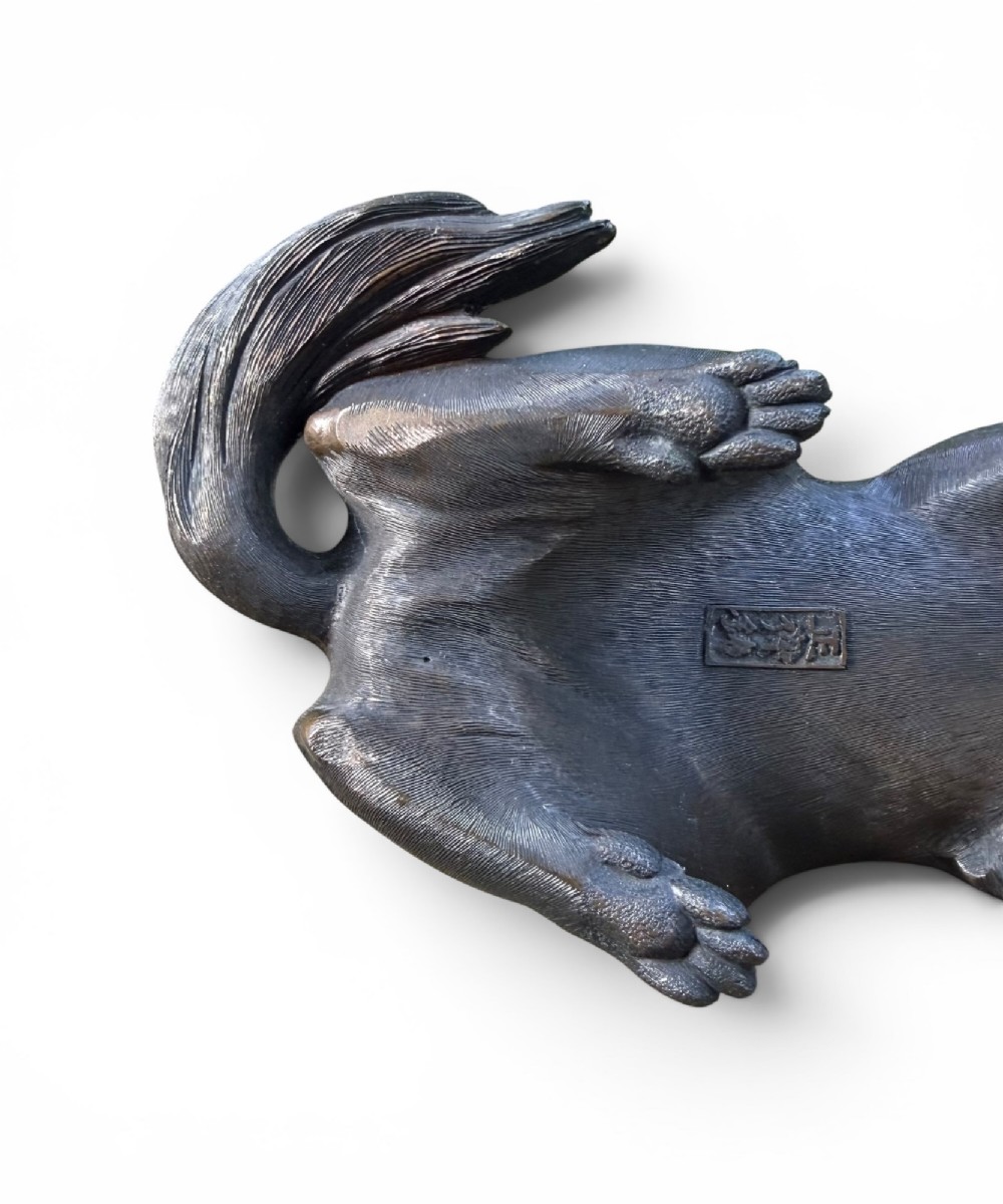 A Japanese bronze sculpture depicting a seated tanuki resting its paw on corn husks, featuring textured fur engraving and a dark patinated finish. View IX