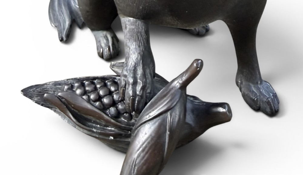 A Japanese bronze sculpture depicting a seated tanuki resting its paw on corn husks, featuring textured fur engraving and a dark patinated finish. View X