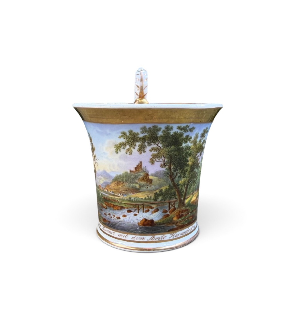 An early nineteenth-century flared porcelain cabinet cup featuring a hand-painted landscape scene with a river, bridge, and hilltop ruins above inscribed German text, finished with a gilded rim and elevated handle.