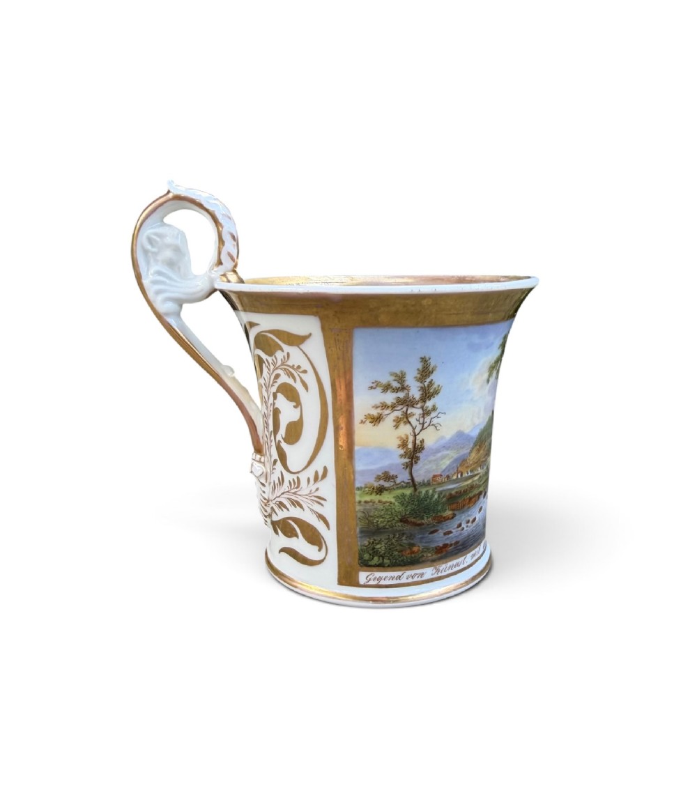 An early nineteenth-century flared porcelain cabinet cup featuring a hand-painted landscape scene with a river, bridge, and hilltop ruins above inscribed German text, finished with a gilded rim and elevated handle. View II