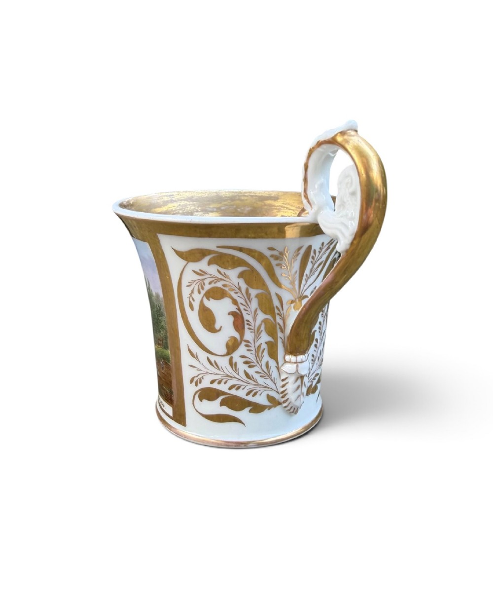 An early nineteenth-century flared porcelain cabinet cup featuring a hand-painted landscape scene with a river, bridge, and hilltop ruins above inscribed German text, finished with a gilded rim and elevated handle. View IV