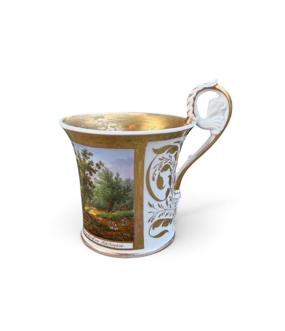 An early nineteenth-century flared porcelain cabinet cup featuring a hand-painted landscape scene with a river, bridge, and hilltop ruins above inscribed German text, finished with a gilded rim and elevated handle. View V