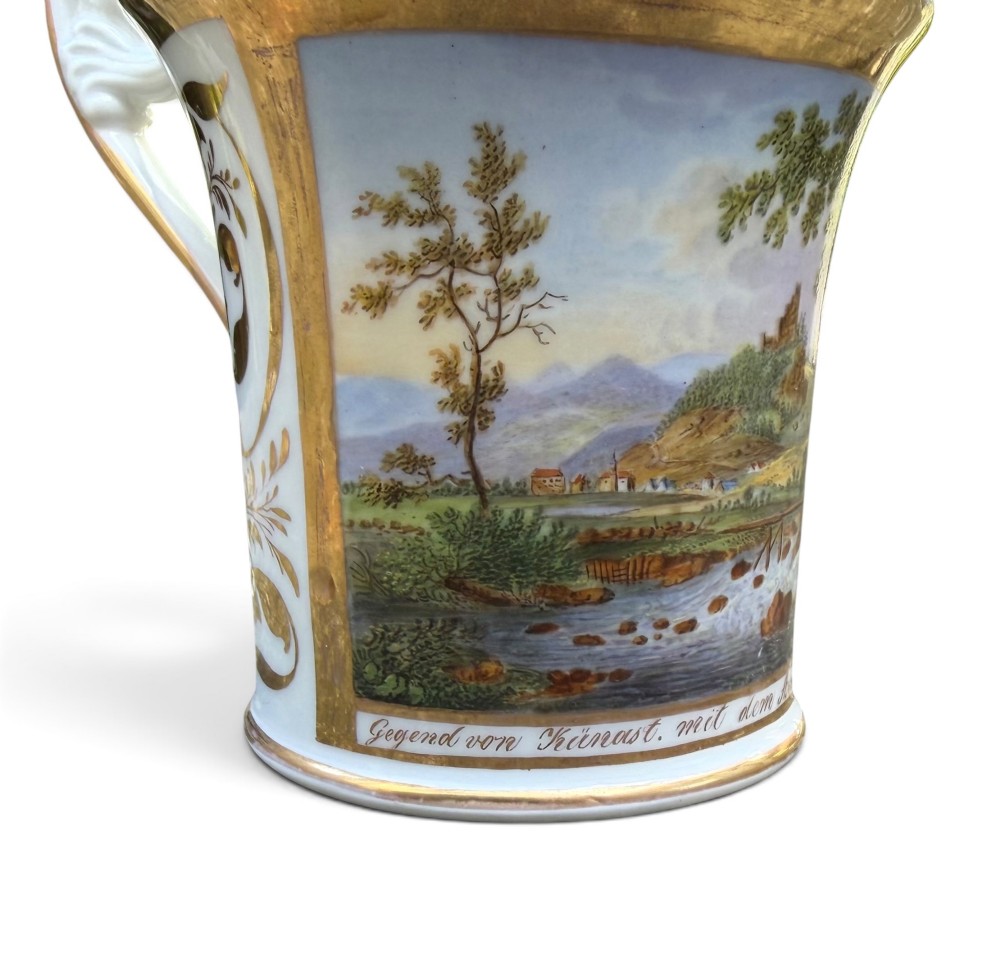 An early nineteenth-century flared porcelain cabinet cup featuring a hand-painted landscape scene with a river, bridge, and hilltop ruins above inscribed German text, finished with a gilded rim and elevated handle. View VIII