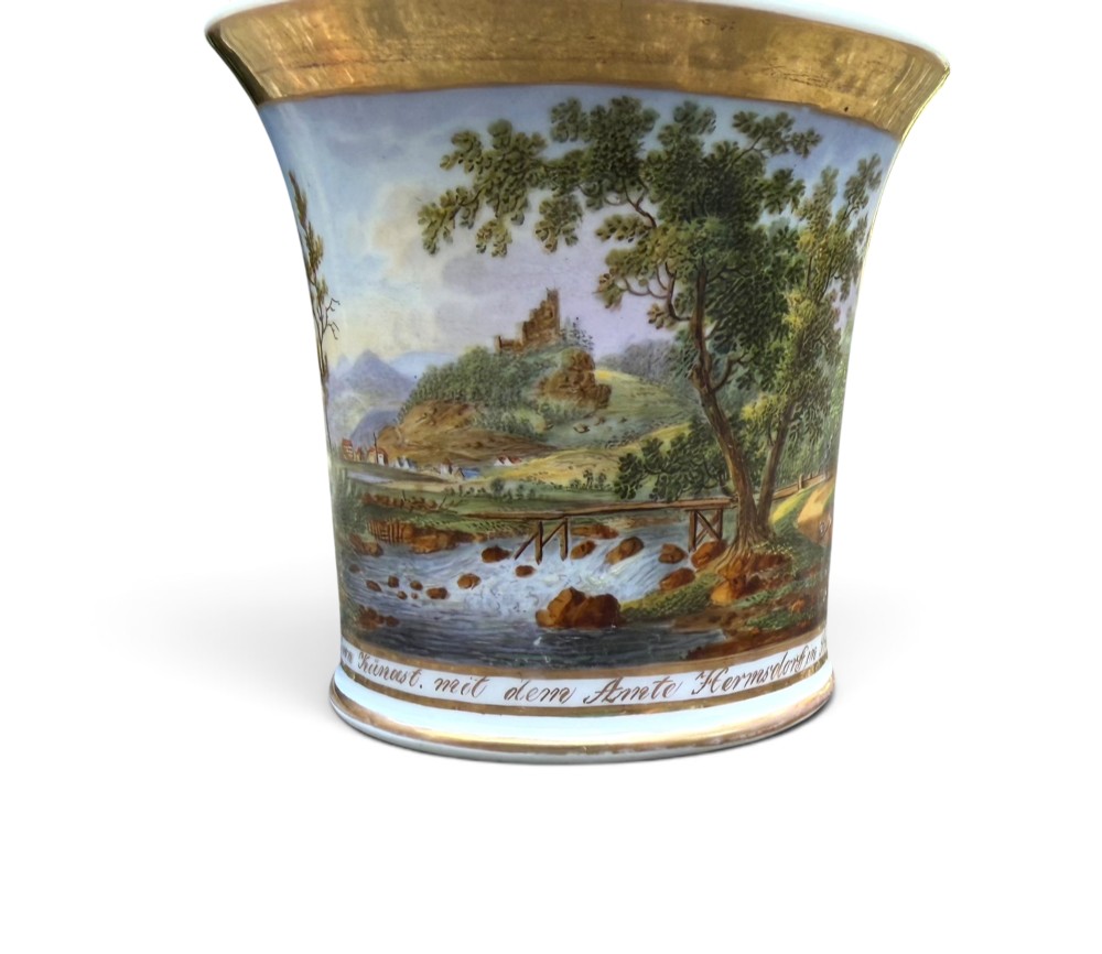 An early nineteenth-century flared porcelain cabinet cup featuring a hand-painted landscape scene with a river, bridge, and hilltop ruins above inscribed German text, finished with a gilded rim and elevated handle. View IX