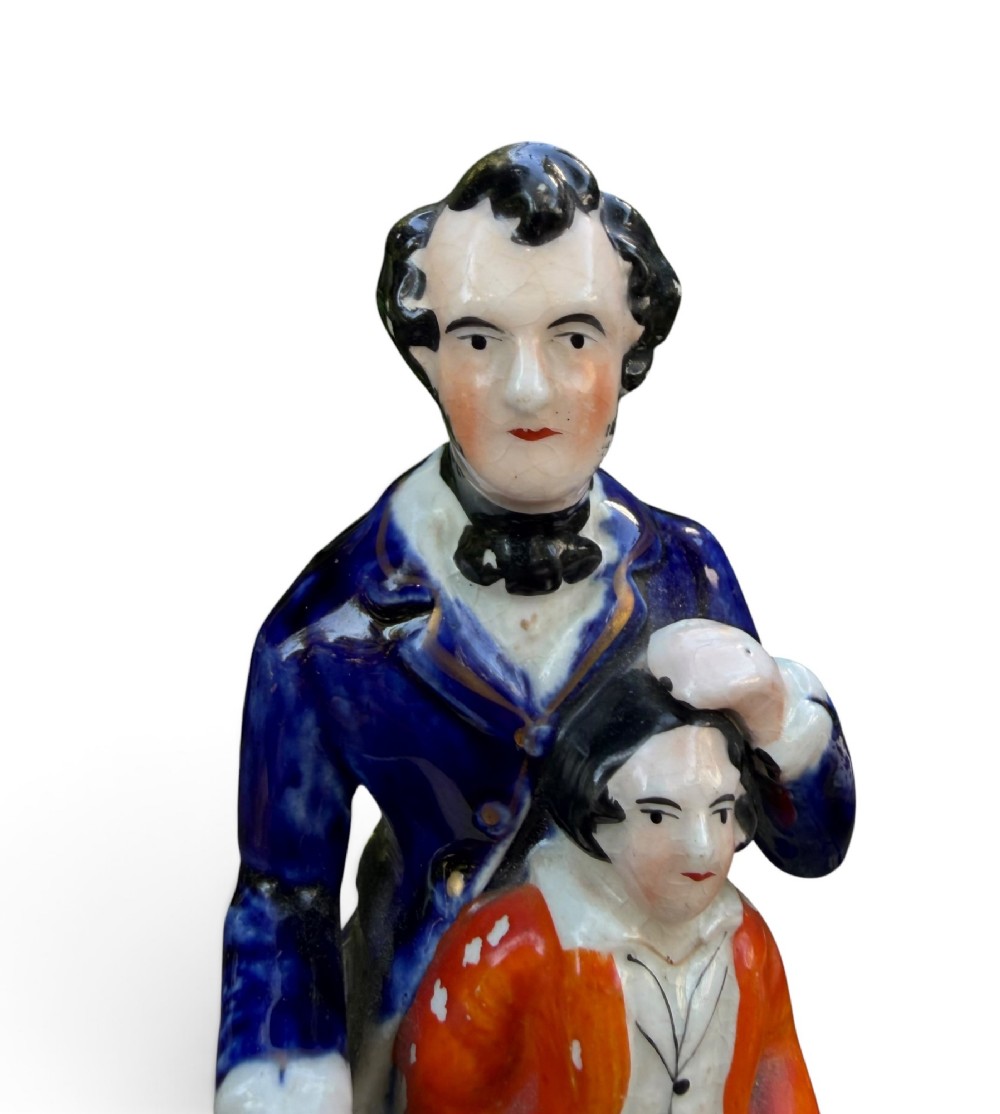 An antique Victorian Staffordshire earthenware figure depicting Lord Shaftesbury in a cobalt blue coat seated with a young child in an orange jacket on a white base. View V