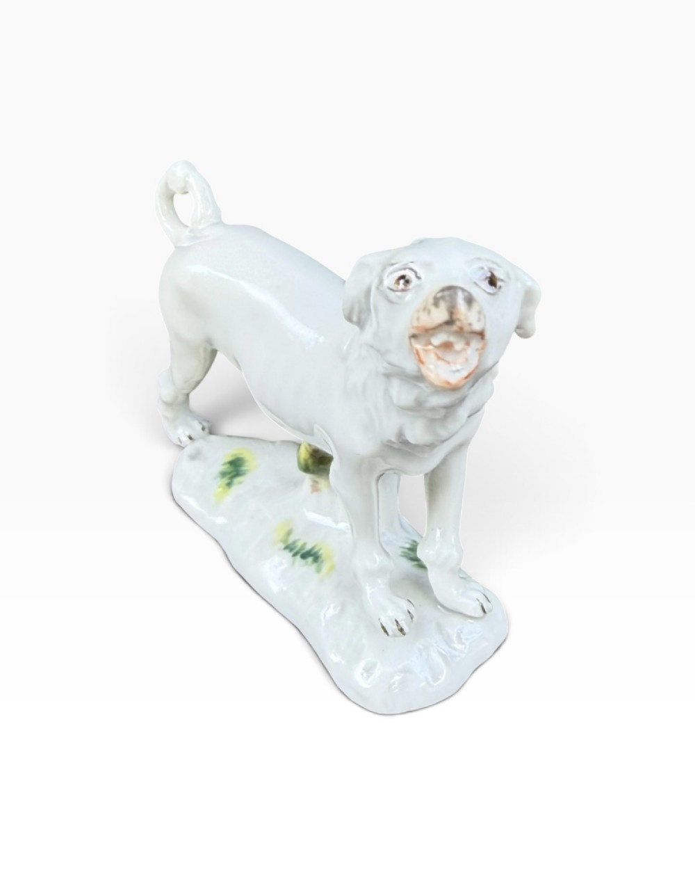 A white glazed Meissen porcelain dog figure standing with an open mouth, curled tail, and a subtle green-painted tree stump base support. View II