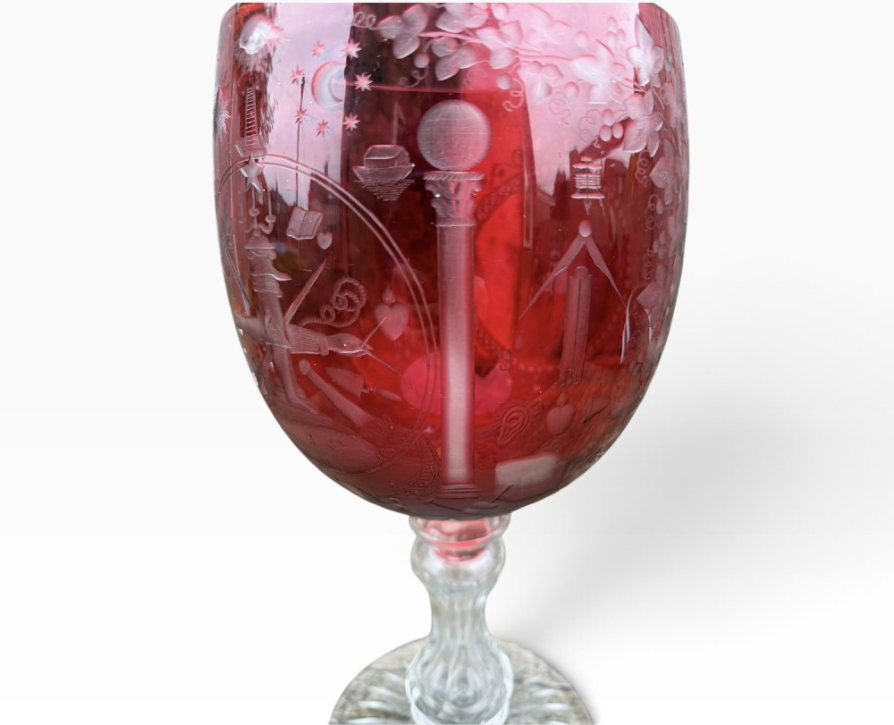 A Victorian cranberry glass goblet with intricately wheel-engraved Masonic symbols on a red bowl, supported by a clear knopped glass stem and a star-cut circular base. View V