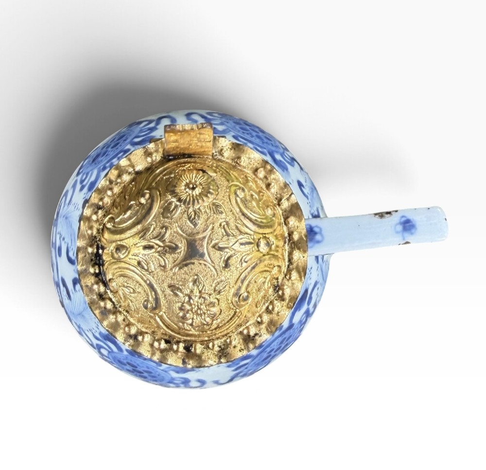 A blue and white Kangxi period Chinese porcelain jug with hand-painted floral decoration, an angular ceramic handle, and a chased gilt-metal lid. View X