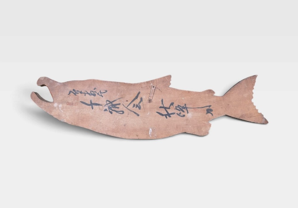 A realistic hand-carved wooden sculpture of a salmon with detailed scales and an open mouth, finished in muted grey and brown tones. View V