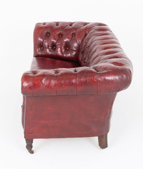 fig 1. An antique Victorian oxblood red leather Chesterfield settee featuring deep buttoned upholstery, rolled arms, hand-tacked studding, and turned wooden legs on brass castors.