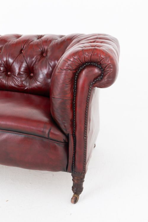 fig 2. An antique Victorian oxblood red leather Chesterfield settee featuring deep buttoned upholstery, rolled arms, hand-tacked studding, and turned wooden legs on brass castors.