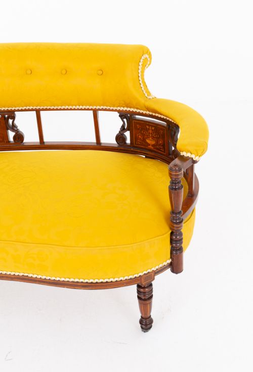 fig 2. An antique curved rosewood settee upholstered in textured yellow fabric, featuring button-backed quilting, turned spindle supports, marquetry inlay panels, and legs on castors.