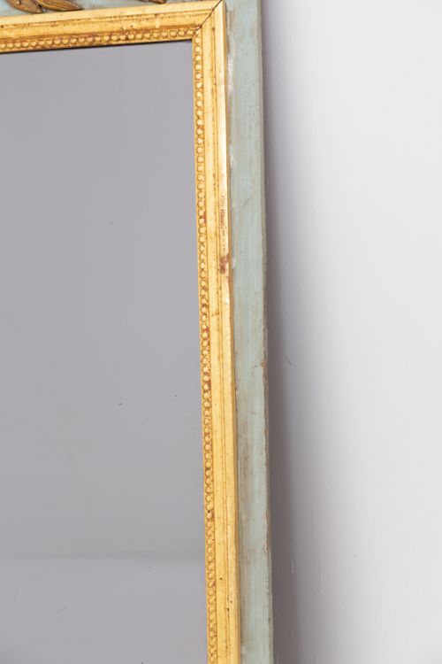 fig 1. A tall French pier mirror featuring a rectangular gilded frame and a detailed carved cresting with a torch and quiver on a light blue wood backing.