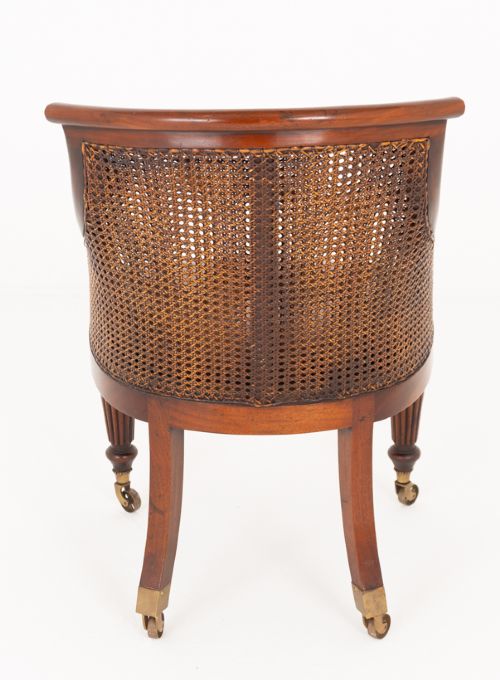 A mahogany Regency Revival desk chair featuring a curved woven cane backrest, a brown leather seat cushion, carved scroll arms, reeded front legs, and brass castors. View V