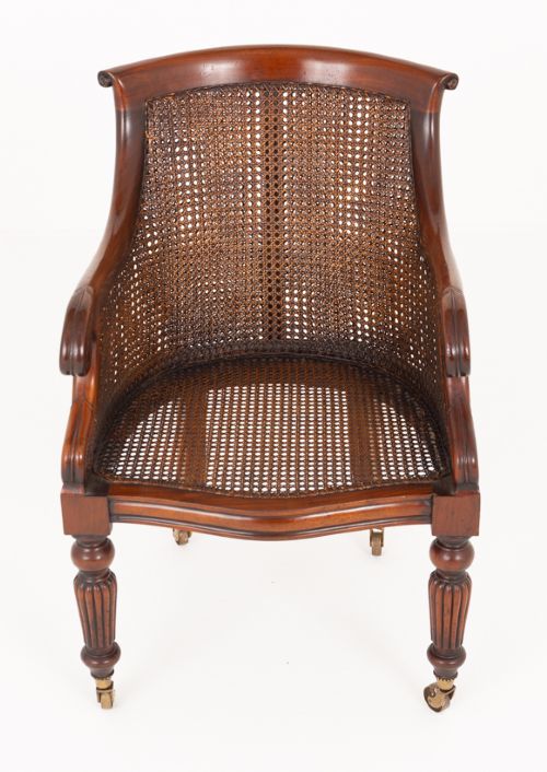 A mahogany Regency Revival desk chair featuring a curved woven cane backrest, a brown leather seat cushion, carved scroll arms, reeded front legs, and brass castors. View VI