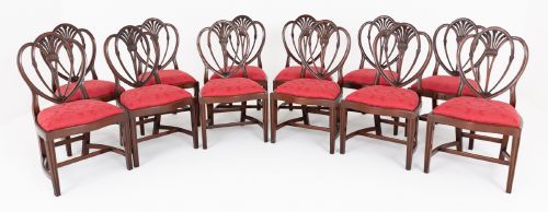 fig 1. A set of twelve mahogany dining chairs with carved Prince of Wales plume backs and red damask fabric seats arranged in a arc.