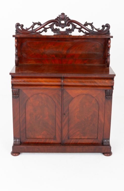 fig 1. A nineteenth-century Victorian mahogany chiffonier featuring a carved crest backboard, a raised shelf with scrolled supports, a cushion drawer, two panelled doors, and bun feet.