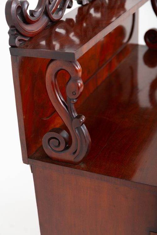 A nineteenth-century Victorian mahogany chiffonier featuring a carved crest backboard, a raised shelf with scrolled supports, a cushion drawer, two panelled doors, and bun feet. View V