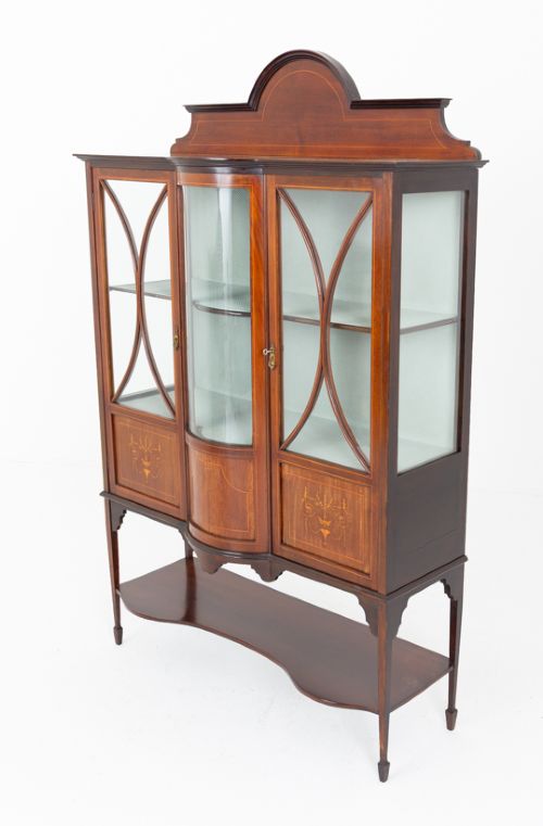 A Sheraton Revival mahogany display cabinet featuring an arched pediment, glass-front doors with geometric glazing bars, marquetry inlay panels, and a lower shelf supported by slender legs. View VI