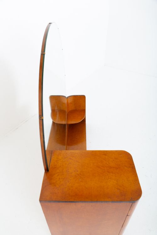 An asymmetric Art Deco dressing table featuring warm birds eye maple veneer, a large circular mirror, a four-drawer stack on the left, and tiered open shelving on the right. View VII