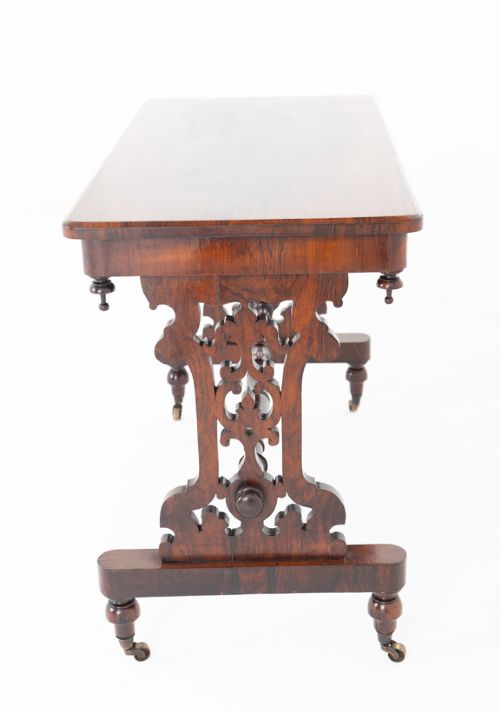 A mid-19th century Victorian rosewood stretcher table featuring a rectangular polished top, openwork pierced end supports, a turned central stretcher, and brass castors. View VI