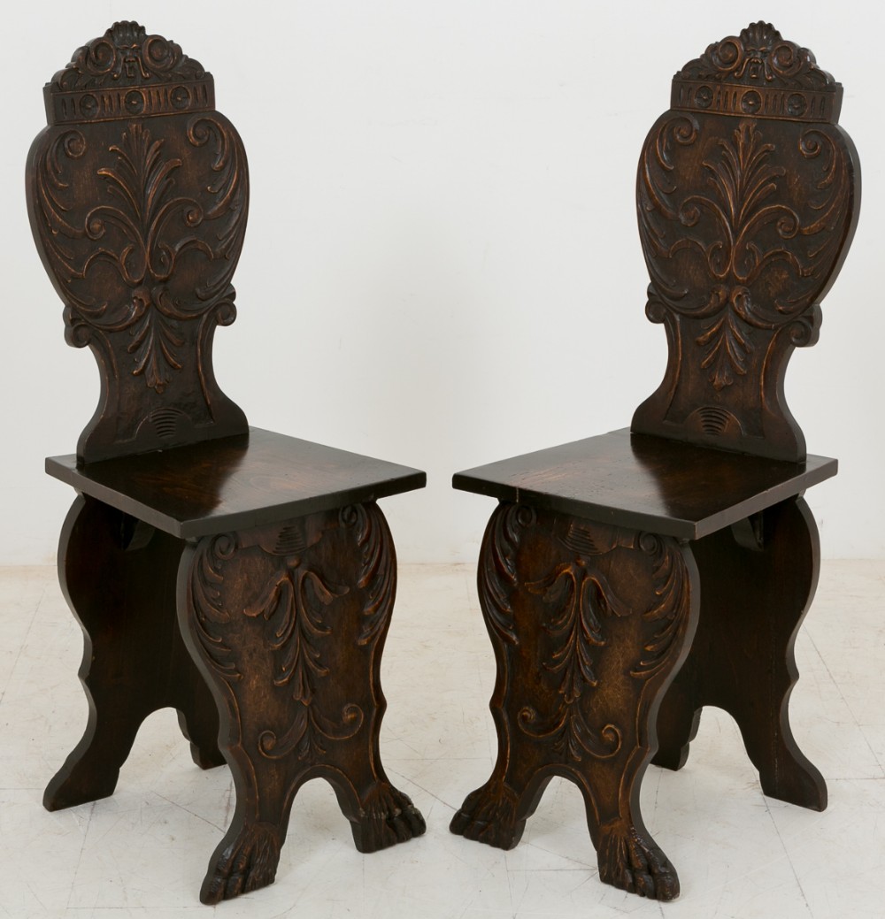 A pair of dark-stained carved limewood Italian hall chairs featuring detailed foliate scrollwork on the backrests and sculpted legs.