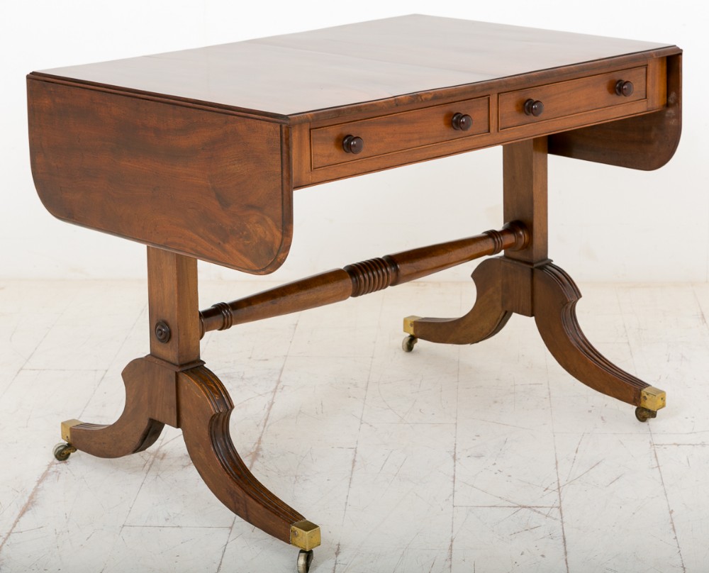 A Regency mahogany sofa table featuring two drop leaves, two shallow drawers with wooden knobs, a turned stretcher, and splayed legs ending in brass castors.