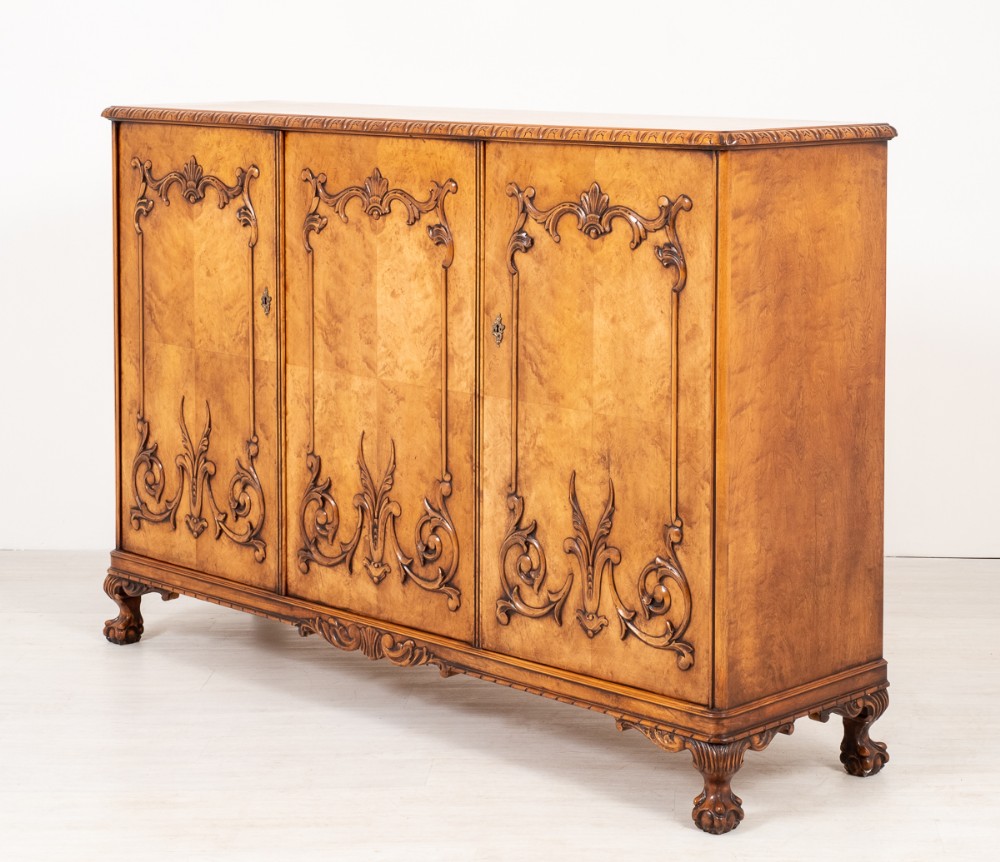 A three-door birds eye maple cabinet featuring applied carved scrollwork on the door panels, a carved lower apron, and claw feet.