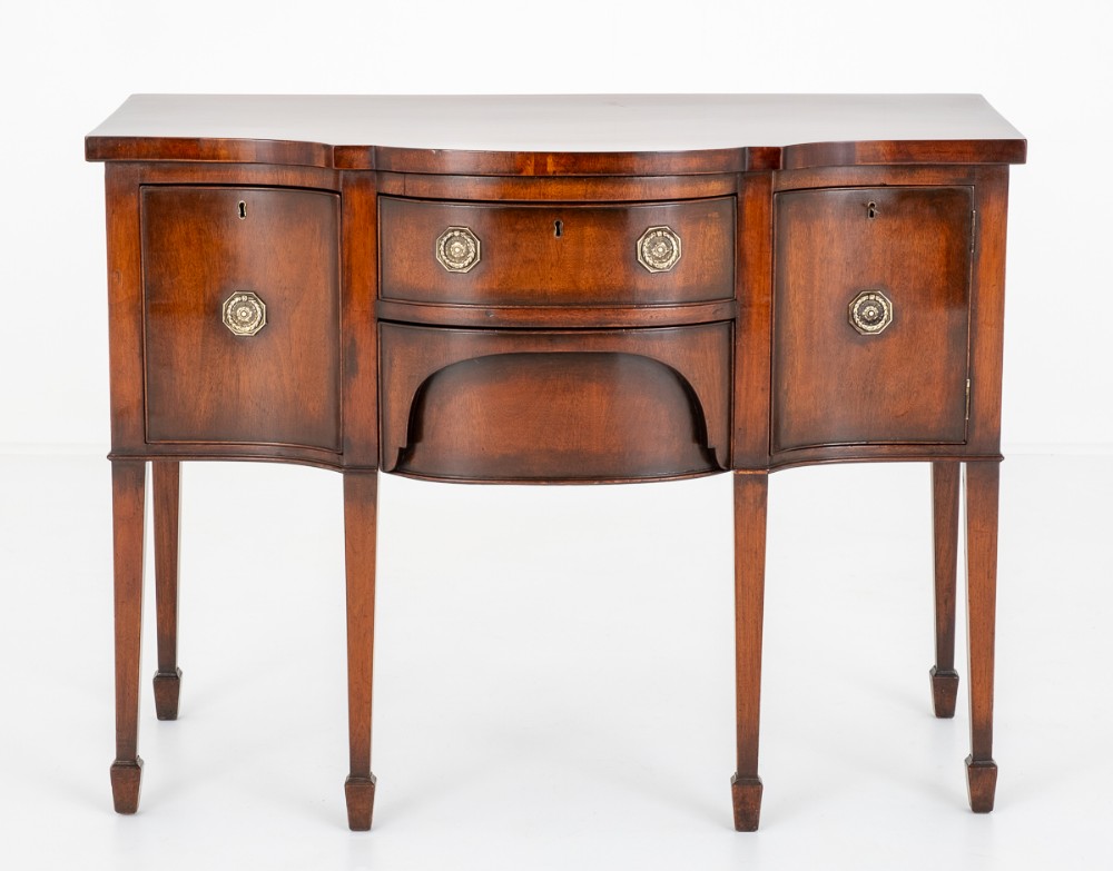 A Regency style polished mahogany sideboard featuring a bow-front silhouette, central drawer, flanking cupboard doors, circular brass hardware, and six square tapered legs.