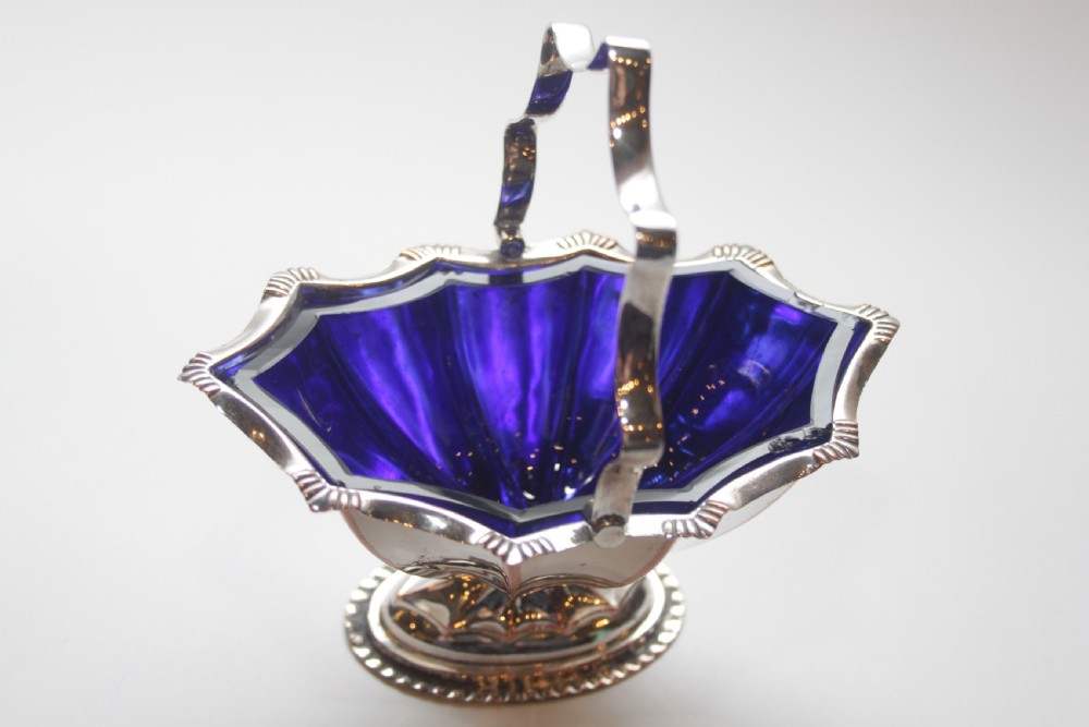 A silver-plated pedestal bon bon dish featuring a scalloped rim, a swing handle, and a cobalt blue glass insert.