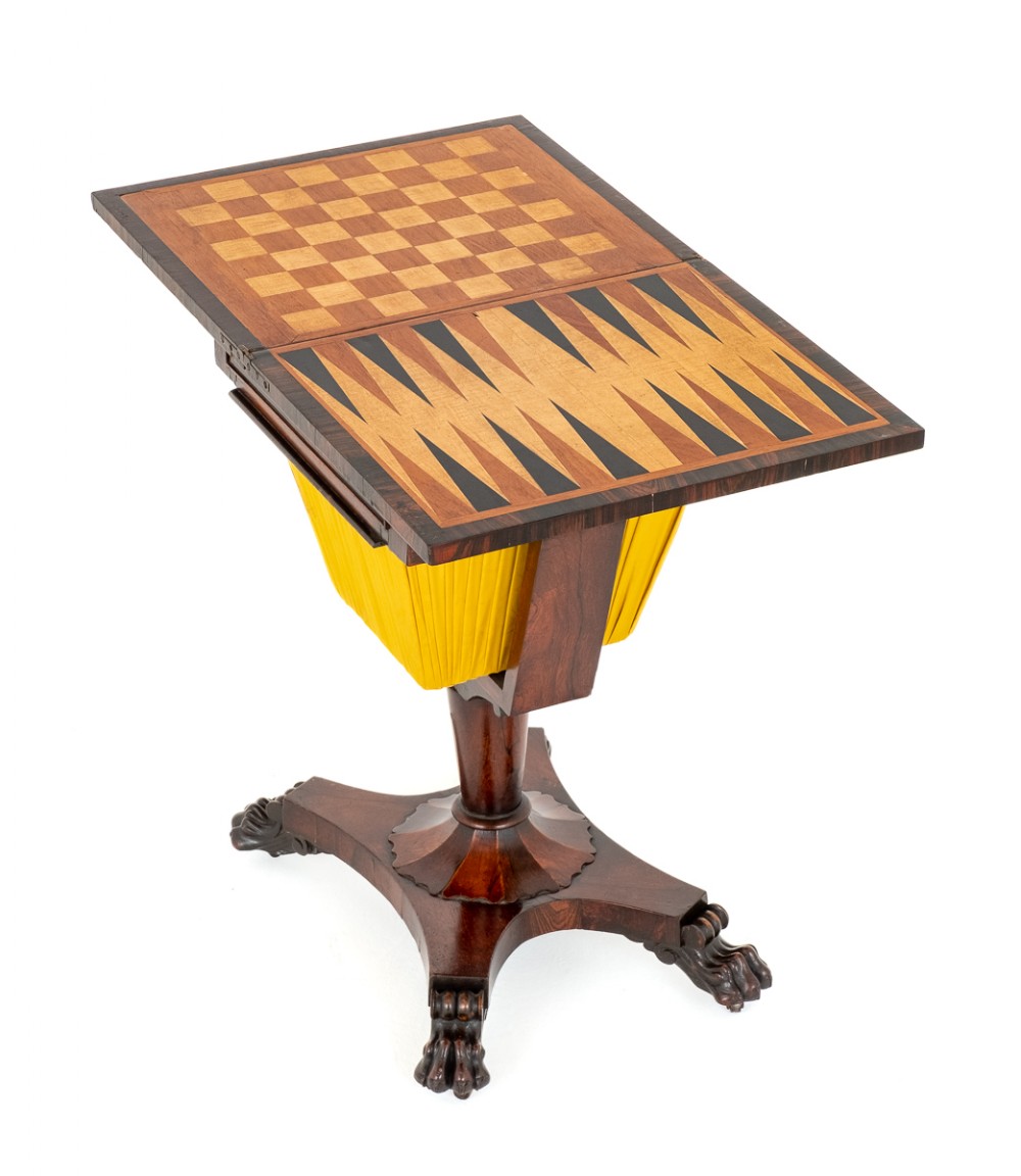 A Regency rosewood games table featuring an inlaid chessboard and backgammon top over a yellow pleated fabric work basket, raised on a pedestal with carved paw feet.