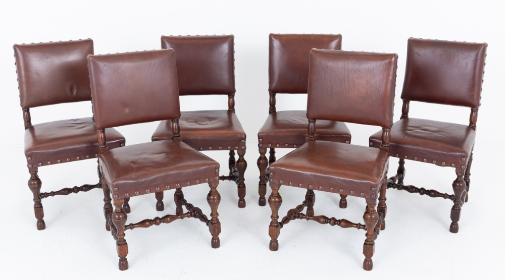 A set of six Tudor Revival oak dining chairs with rectangular dark brown leather seats and backs secured by metal studs.