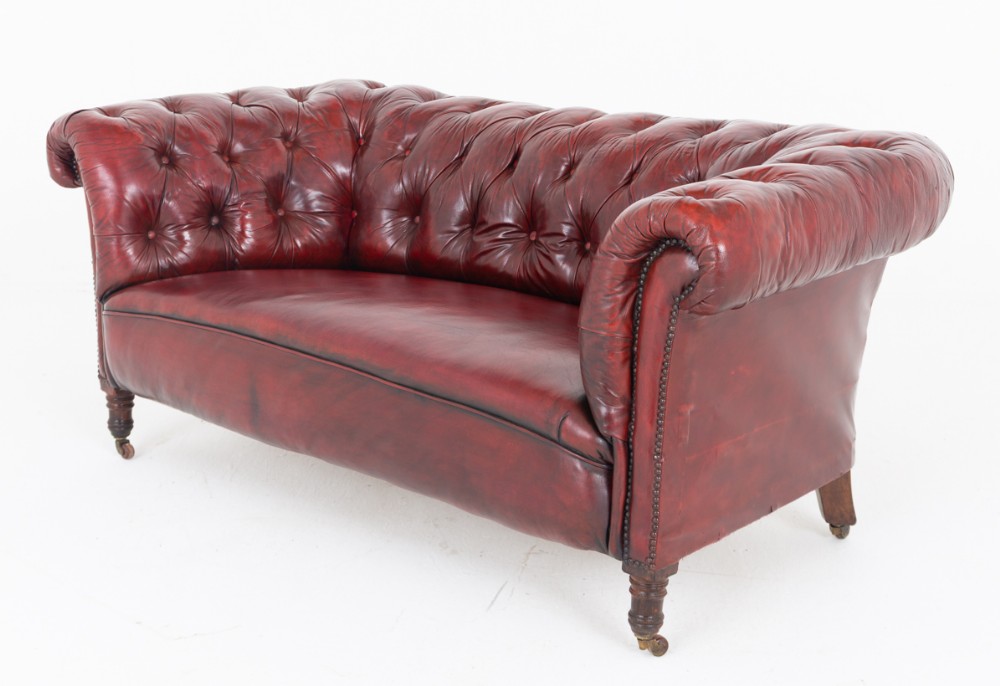 An antique Victorian oxblood red leather Chesterfield settee featuring deep buttoned upholstery, rolled arms, hand-tacked studding, and turned wooden legs on brass castors.
