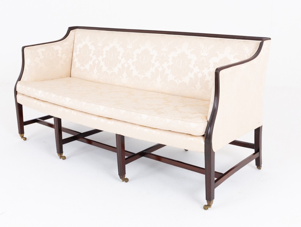 A Georgian mahogany settee upholstered in cream patterned damask fabric with dark straight legs, cross-stretchers, and brass castors.