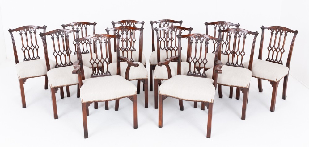 A set of twelve carved mahogany dining chairs in the Gothic Revival style, featuring ten side chairs and two carvers with neutral upholstered seats.