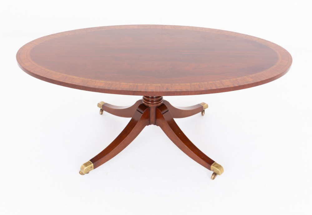 An oval mahogany coffee table featuring a crossbanded inlaid top, turned central pedestal, four splayed legs, and brass cap castors.