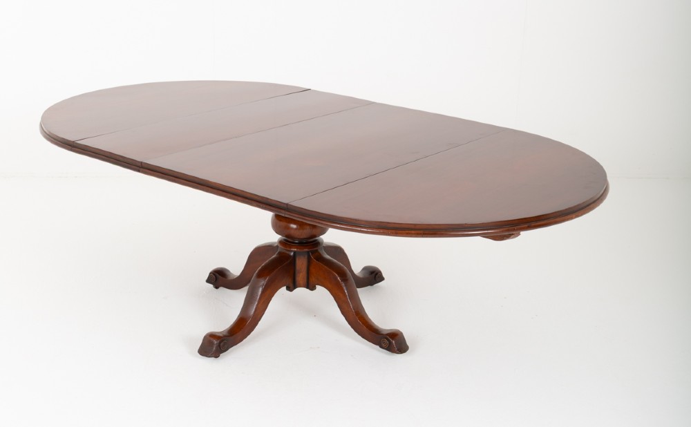 An antique Victorian mahogany extending dining table featuring an oval top, removable centre leaf, and a carved central pedestal base with splayed legs.