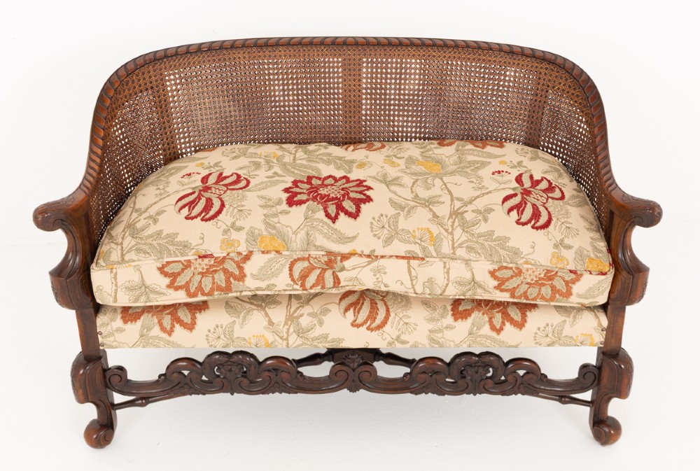 A three-piece Carolean style seating suite comprising a settee and two matching armchairs with woven cane backrests, carved scroll stretchers, and floral fabric seat cushions. View IV
