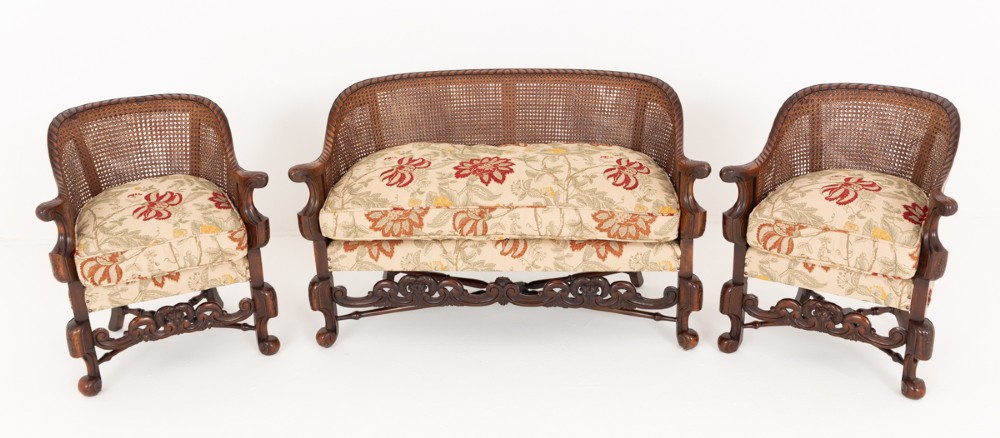 A three-piece Carolean style seating suite comprising a settee and two matching armchairs with woven cane backrests, carved scroll stretchers, and floral fabric seat cushions.