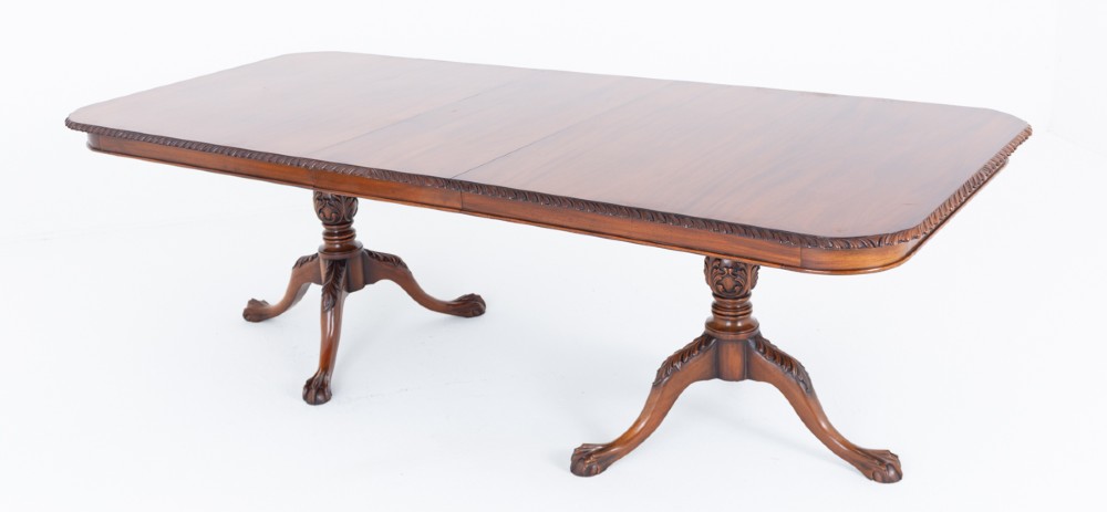 A Regency Revival twin-pedestal mahogany dining table featuring carved edging, turned support pillars, and splayed legs ending in carved feet.