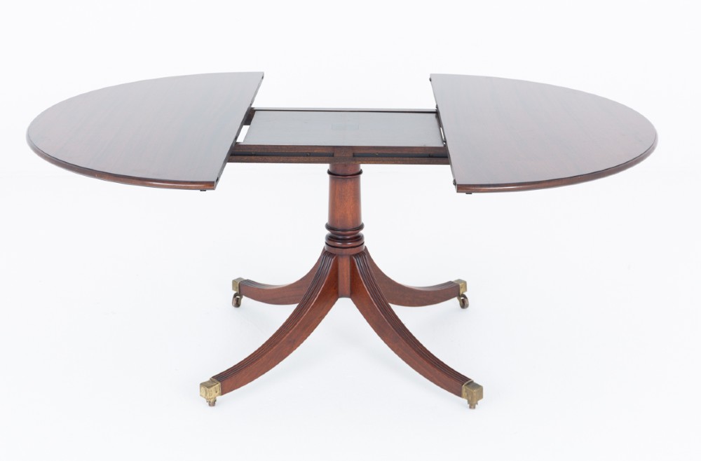 An extendable Regency Revival oval mahogany dining table with a central turned pedestal support, four reeded swept legs, and brass castors. View V
