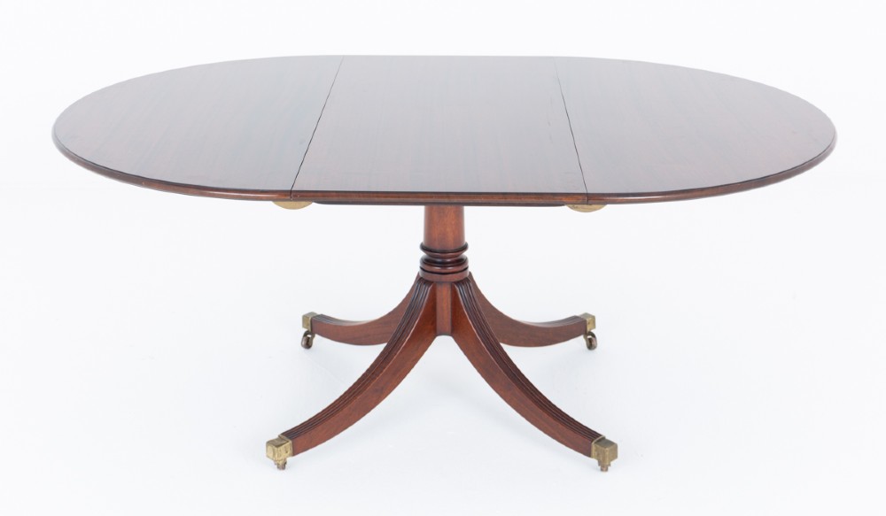 An extendable Regency Revival oval mahogany dining table with a central turned pedestal support, four reeded swept legs, and brass castors. View VI