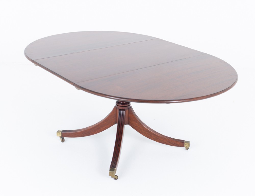 An extendable Regency Revival oval mahogany dining table with a central turned pedestal support, four reeded swept legs, and brass castors.