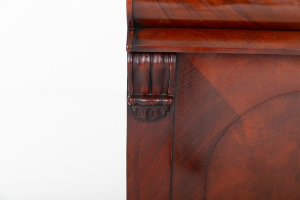 A nineteenth-century Victorian mahogany chiffonier featuring a carved crest backboard, a raised shelf with scrolled supports, a cushion drawer, two panelled doors, and bun feet. View VI
