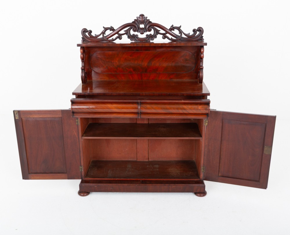 A nineteenth-century Victorian mahogany chiffonier featuring a carved crest backboard, a raised shelf with scrolled supports, a cushion drawer, two panelled doors, and bun feet. View VII